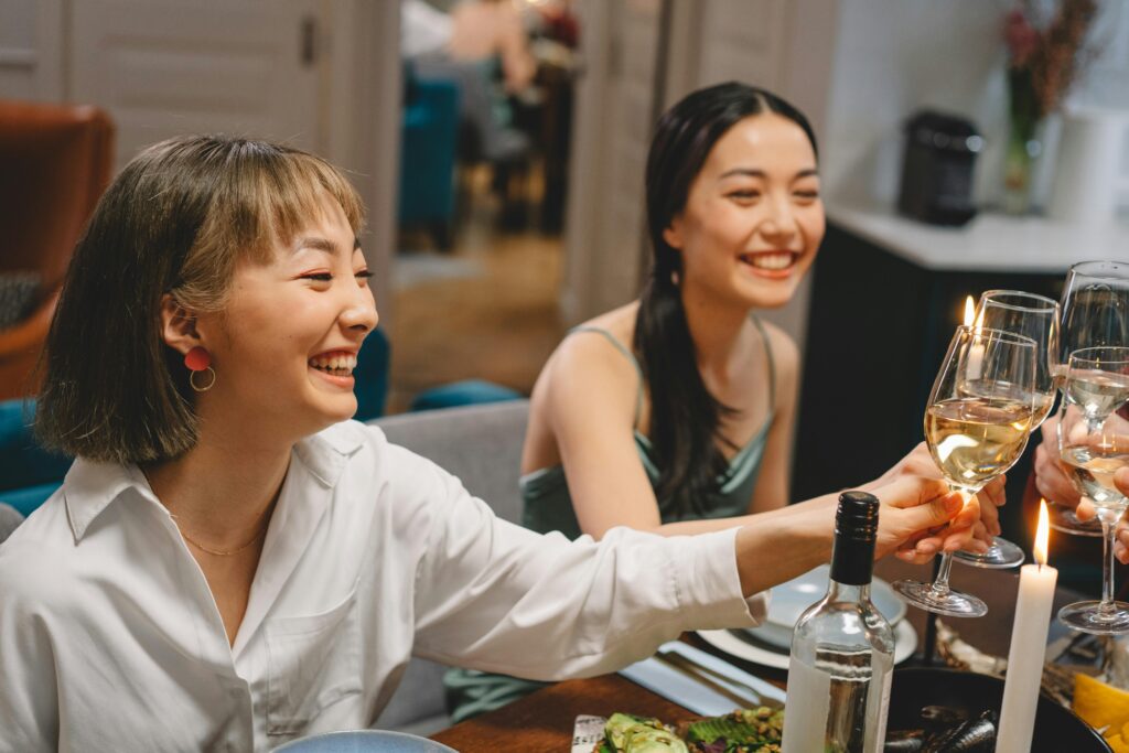 Two women enjoying a lively dinner party, clinking glasses of white wine in a cozy setting.